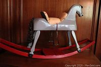 Side view of the gray hand carved wooden rocking horse showing red rocker base and cream leather saddle with metal stirrups and hair mane and tail.