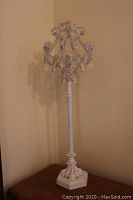 Full view of the white shabby chic tall table lamp on a wooden surface showing tall turned stand and crystal decorated top.