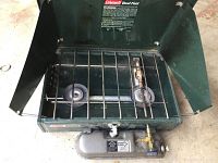Top view of the Coleman dual fuel camp stove showing two burners, removable chrome-plated grate, and fuel tank with valve and pump.