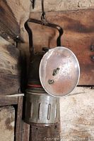 Miner's carbide lamp with round metal reflector and handle, showing patina and signs of use, hanging on a hook against wooden background.