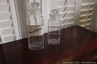 Two clear antique glass apothecary jars with stopper lids on a dark wood surface, with plantation shutters in background. The larger jar features a vintage label 'PV.con.INSECT.'