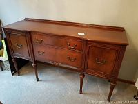 Solid wood vintage sideboard front and side angles showing two drawers in the center flanked by cupboards with paneled doors and decorative handles, standing on fluted legs.