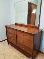 Front and angled side view of wooden dresser with six drawers and attached large mirror. Brass handles on drawers with wing design.