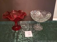 Two glass fruit bowls, one clear and one red, on a green tablecloth against a brown background