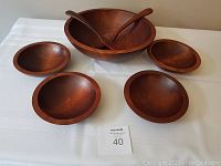 Large wooden salad bowl with two wooden serving utensils and four matching small wooden bowls arranged on a white tablecloth.