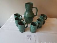 Full set display of one large green pitcher and seven matching mugs on a white surface.