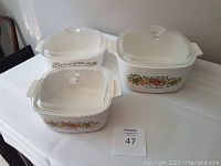 Three Corningware baking dishes with lids placed on a white tablecloth near a wall corner, each featuring vegetable floral pattern on the side.