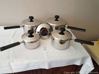 Photo showing four stainless steel cooking pots with black handles, each paired with a lid with black knob, arranged on a white tablecloth.