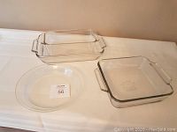 Three clear glass Pyrex baking dishes displayed on a table cloth against a wall: two rectangular casserole dishes with handles and one round dish.