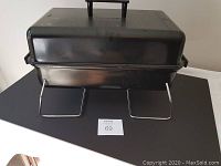 Closed portable propane BBQ grill showing black metal housing and foldable metal legs.