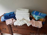 Wide shot of all towels stacked on wooden dresser: white bath towels center, blue bath towels left, beach towels right, assorted hand and face cloths front edges.