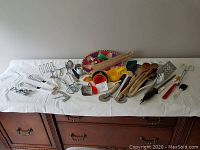 Full view of the kitchen utensil assortment on a white cloth on top of wooden furniture: multiple utensils including whisks, peelers, wooden spoons, measuring spoons, and a colorful woven basket
