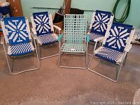 Five folding lawn chairs arranged on floor: four with blue and white macrame weaving and one teal webbed chair in center.