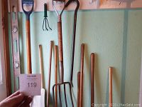 Photo of miscellaneous garden tools including a level, shovels, hand cultivator, and wooden handles around a green wall.