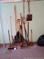 Photo shows a lineup of various garden tools standing against a wall including rakes, shovels, and brooms.
