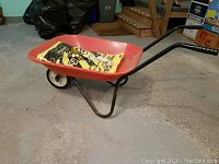 Small red metal wheelbarrow from side angle showing black metal frame, single rubber wheel and black handles, with a bag of salt inside