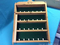 Front view showing four horizontal rows holding white golf ball tees with green felt backing, wooden frame, and a ruler on the left side.