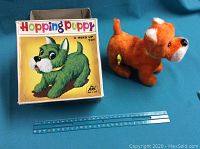 Original orange plush hopping puppy toy with yellow wind-up key, shown beside a ruler for size comparison, with original colorful box featuring a green puppy illustration.