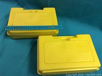 Two yellow LEGO storage cases shown closed and stacked on top of each other on a blue surface with a ruler across one case for scale.