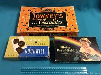 Photo shows three vintage chocolate boxes: Lowney's Chocolates box in orange, Ganong's Goodwill Chocolate box in blue with chocolate pieces, and Mott's Pot of Gold Chocolate box with image of woman and rainbow. Boxes show wear and corner damage.