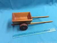 Top-down angled photo of small wooden box cart with two black wheels and long handles resting on blue surface with a ruler for scale.