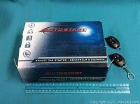 Closed blue AutoStart branded box with 2 black remote car key fobs beside it on a blue background with a ruler.