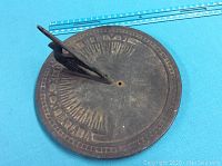 Top-down view of a round metal sundial showing Roman numerals and a sharp triangular gnomon casting a shadow. A blue measuring ruler provides scale.