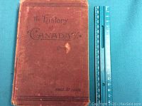 Front cover of a reddish-brown hardcover book titled 'The History of Canada' with decorative elements and a marked price of 50 cents. The book is measured next to a blue ruler to show size.