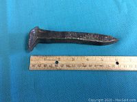 Close-up photo showing one vintage metal railroad spike placed next to a ruler for scale, approximately 6 inches long, with worn surface and flat head typical of railroad spikes.