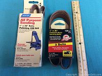 Two packs of sanding belts side by side with a ruler for scale. Left pack is Norton All Purpose Polishing 320 grit, right pack is 3-pack Premium Resin 120 grit belts.
