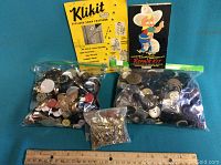 Photo showing two plastic bags filled with assorted buttons, a small bag with metal pins, and two vintage packaged kits — Klikiit snap fasteners and Repu Repair Kit.