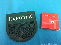 Photo of black rubber Export A store change pad and unopened du Maurier cigarette pack box side by side on blue surface.