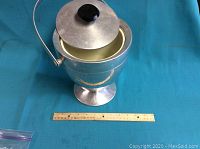 Full view of the silver-tone metal wine cooler showing pedestal base, hinged lid with black knob, and carrying handle. Ruler for size reference.