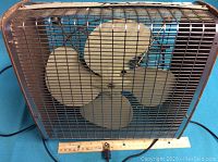 Front view of the Torcan metal box fan showing four blades and metal protective grill with ruler for size reference.