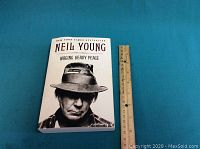 Paperback book "Waging Heavy Peace" by Neil Young on a blue surface alongside a ruler for size reference.