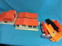 Plastic building sections with orange roofs and white walls, a ruler for scale, and a pile of orange and black track pieces.