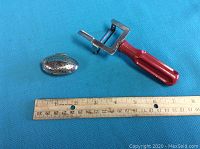 Red handled vegetable peeler and perforated metal tea bag holder with ruler showing size