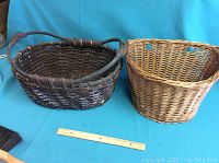Two wicker baskets shown side by side on a flat surface with a blue backdrop. A ruler and partial broom visible for scale and context.