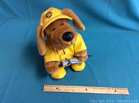 Brown plush Nova Scotia hound in yellow raincoat and hat holding umbrella on blue backdrop with ruler for size reference.