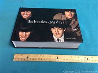 Closed hardcover book with black cover showing painted portraits of The Beatles members and title 'the beatles 365 days simon wells'. Ruler placed below for scale showing approximately 9 inch width.