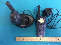 Front view showing one walkie talkie, charging base, and power adapter against blue background and wooden ruler for scale.