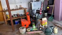 Photo shows orange snow blower, two ceramic flowerpots, metal and plastic watering cans, garden flag on a pole, spade shovel, and various garden watering supplies on garage floor