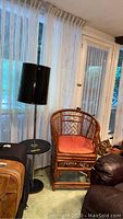 Black floor lamp, rattan armchair with orange cushion, small black glass topped side table in room corner with sheer curtains