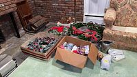 Photo showing multiple Christmas wreaths including one with a large red bow, cardboard boxes with additional decorations and a silver pitcher beside a floral sofa.