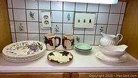 Kitchen countertop showing all items in the lot including the Wedgwood dinner plates, Franciscan mugs and plates, Johnson Brothers pitcher and bowl, in front of a tiled wall.