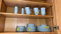 Cup and plate sets arranged on wooden shelves inside a cabinet. Top shelf has 6 cups, bottom shelf has stacks of plates and saucers with yellow tags.