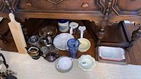 Photo showing a variety of glass, ceramic, and metal kitchen and serving items placed on a table. Includes teapots, bowls, pitchers, and baking dishes under a wooden dresser.