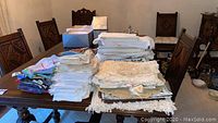 Photo of multiple stacks of folded tablecloths, linens, and placemats on a wooden dining table with chairs in the background.