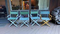 Four director chairs lined up on an outdoor deck near a glass door. Three chairs have blue painted wood frames and one has a natural wood finish. All have greenish-blue cloth seats and backs and show outdoor wear.