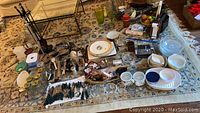 Wide view of assorted kitchen cutlery, dishes, mugs, glass and ceramic bowls, plastic containers, and fireplace accessories laid out on a rug.